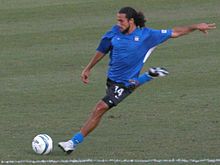 Dwayne De Rosario, vestido con una camiseta azul, se prepara para golpear un balón de fútbol en el suelo con el pie derecho.