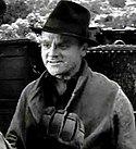 Head and shoulders shot of Cagney, wearing black fedora and smiling slightly, scenery in the background