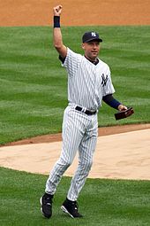 Мужчина в белой бейсбольной форме в темно-синюю полоску поднимает правую руку в воздух, держа коробку в левой руке.