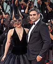 A picture of a couple embracing each other. The woman standing on the left is wearing a black dress paired with a black headdress and a diamond necklace, and the man standing on the right is wearing a black tuxedo, a white shirt and a black bow tie.