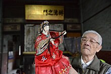 A female hand-puppet wearing a red Taiwanese dress is held up at head-height, worked by an old man in Western clothing with delicate glasses and short white hair.