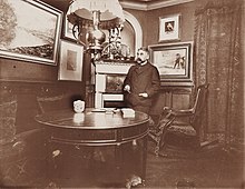 Photographie sépia d'un homme se tenant debout près d'une table dans un appartement bourgeois.