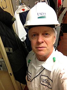 Ted Conover wears a hairnet, a USDA hardhat, and a USDA inspector uniform while standing in front of a dressing room locker.