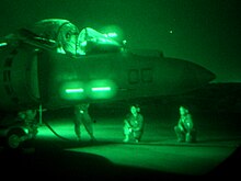 Mostly dark with green hue, this is a night-vision of jet aircraft and several mechanics kneeling close by. The aircraft's canopy is open.