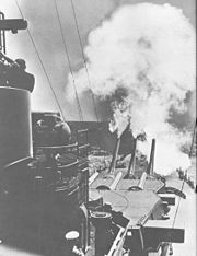 Six large guns in two turrets are aimed directly forward; the far trio are elevated extremely high and have a large amount of smoke emanating from them