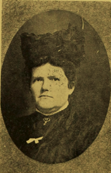 B&W portrait photo of a woman wearing a dark blouse with a white ribbon pinned on it, as well as a dark hat.
