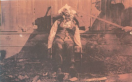 Body of a lynched black male, propped up in a rocking chair for a photograph, circa 1900. Paint has been applied to his face, circular disks glued to his cheeks, cotton glued to his face and head, while a rod props up the victim's head.