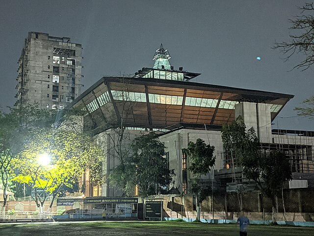 Nighttime photo of a large, square concrete building topped by an inverted trapezoidal section (wider at the top than the bottom) ringed by a row of windows, and a much smaller, almost all-glass rectangular room atop that, in turned topped with a narrow glass pyramid partly surrounded by decorative metal bars. In the foreground are several trees and a sports field, and in the background to the left there's a cold gray, rectangular high-rise office building