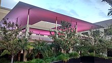 A roofed structure, open to the elements, with a large skylight. The structure is surrounded by trees. Artificial lighting makes the underside of the roof pink, in contrast with the light blue of the sky at dusk.