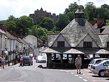 Un pequeño edificio de un solo piso con un techo en forma de pirámide, al costado de una carretera bordeada de edificios.  Algunos coches pequeños privados visibles.  Árboles en la distancia con el horizonte del castillo de Dunster.