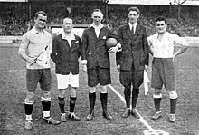 Cinq personnes debout au milieu d'un terrain de football : les officiels au centre et deux joueurs de football sur les cotés.