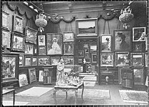 Vue d'une partie d'une salle de la galerie Henry Vasnier. Sur les murs sont accrochés des tableaux, d'autres sont posés à même le sol, contre eux. Au centre se trouve une table sur laquelle sont posés des sculptures et des objets d'art. Des livres sont rangés dans des meubles devant les cimaises. Des tapis recouvrent le sol. Une porte ouverte laisse entre voir la salle suivante.