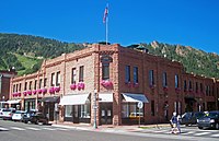 Un bâtiment en pierre brun clair de deux étages au coin d'une rue, avec une entrée étroite face à la caméra au centre de l'image.  Il est éclairé par le soleil de gauche.  Il y a des devantures de magasins des deux côtés au niveau de la rue;  toutes les fenêtres du deuxième étage.  Au-dessus de l'entrée principale se trouve un auvent avec le mot "Dior" dessus, et au-dessus de la fenêtre du deuxième étage une plaque noire semi-circulaire avec "Brand Building, 1891, Aspen, Colorado" en lettres dorées.  Un drapeau américain est sur un poteau au-dessus.  En arrière-plan se trouve une ligne de crête rocheuse et boisée avec quelques zones défrichées.