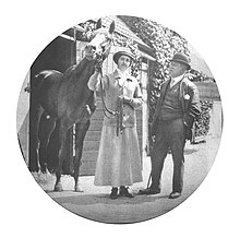 A woman in a long skirt and jockey's cap holding the reins a horse on her right with a man in a suit standing to her left.