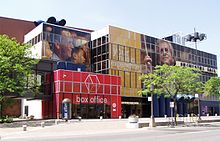 Modern building, red entrance labeled BOX OFFICE, in front of yellow and blue segments. Trees planted in front. Advertising has images of people's heads painted on building.