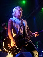 English guitarist Peter Hook performing live in front of a dark background, wearing a white tank top and jeans.