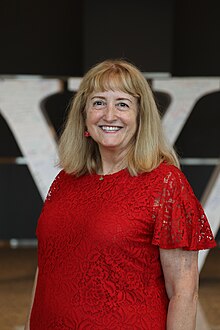 Rosie Stephenson-Goodknight in front of the Cardboard W in Wikimania 2023. She is a middle-aged white woman with blonde hair wearing a red dress.