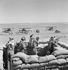 photographie en noir et blanc montrant un groupe de soldat dans une fortification en sacs de sable avec des avions biplan stationnés à l’arrière-plan