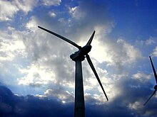 Photographie couleur. Extrémité d'une éolienne avec trois pales se détachant dans un ciel nuageux.