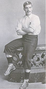 אדם עם שפם כהה, בחולצת כדורגל בהירה, מכנסיים קצרים כהים ארוכים וכובע, יושב על קצה מעקה אבן