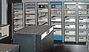 A room with a black box in the foreground and six control cabinets with space for five to six racks each. Most, but not all, of the cabinets are filled with white boxes.