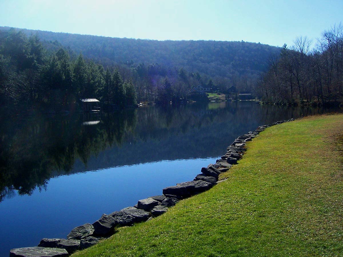 A small lake with a smooth surface reflecting the blue sky above and a hill beyond it with some buildings along the far shore. In the foreground， at lower right， the edge is marked by a stone retaining wall