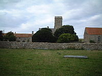 Square stone tower and red roofed buildings behind a stone wall and partially obscured by trees.
