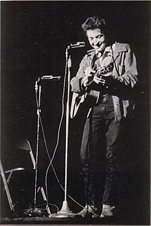 Dylan with his guitar onstage, laughing and looking downwards.