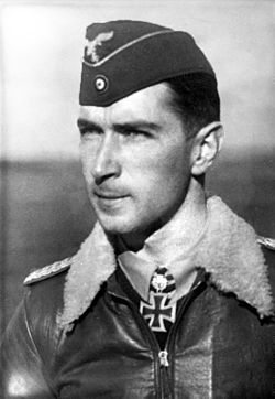 Black-and-white photograph showing a young man in semi-profile, his face pointing in a direction to the left of the camera. The top of his head is covered by a field cap with an eagle emblem and a small concentric-circles motif at the front. He wears a pilot's leather jacket with a fur collar resting on his shoulders, with the central front zip closed to within 2 or 3 inches of his shirt collar, and a Knight's Cross of the Iron Cross with Oak Leaves visible above the zip at the front of his shirt collar. His hair is dark and short, his nose is long and straight, and his facial expression is a determined and confident smile; his eyes gaze into the distance to the left of the camera.