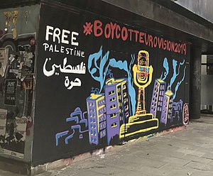 A painted mural on a wall on a street in Girona, Spain: the Eurovision trophy appears covered in barbed wire surrounded by tower blocks, with the words "#BoycottEurovision2019" above, and "Free Palestine" in English and Arabic to the top left