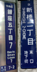 Two "chōmei-name plates (町名板)" are with Romaji for people unable to read the Japanese. (L) - A plate in standard style in larger cities. The letters on the plate indicates from the top Chuō Ward (中央区, Chuō-ku) and Block 7, 5th Neighborhood (銀座五丁目7, Ginza go-chōme nana). And at the very bottom, 7-2, stands for Block 7, Number (banchi) 2. Pictured on the Ginza 4 chōme koban police box at the Ginza 4 chōme crossing, on Ginza main street facing to Wakō. (R) - Pictured is the one without any banchi numbers at the 2nd block in Shimbashi  (新橋二丁目, Shimbashi ni-chōme). In Japanese writing at the bottom it reads you are in the underground city at the east gate (東口地下街, Higashi-guchi chikagai), but the name for Shimbashi station is not indeicated.