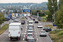 Véhicules reculés sur une chaussée divisée vue d'en haut.  Au premier plan se trouve un feu de circulation ;  il y a des panneaux bleus en allemand plus loin sur la route alors qu'elle se rétrécit.  À l'arrière se trouve une colline développée, partiellement masquée par une brume bleuâtre