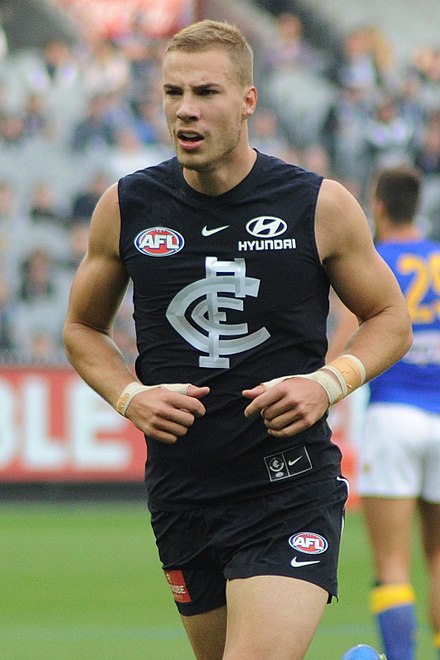 A man with close-cut light brown hair in a sleeveless navy guernsey and shorts