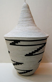 Photograph depicting a bowl shaped off-white woven basket with tall conical lid and black zigzag pattern