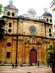 Facade of a yellow stone church with two towers. Above the main entrance there is a human sculpture and higher up a clock.