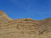 Photo prise depuis le désert de Judée, en contre-plongé, au pied d’une falaise rocheuse, avec un ciel de jour dégagé laissant apparaître la lune
