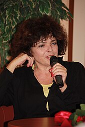 Katarzyna Grochola hat dunkles langes lockiges Haar. Sie sitzt an einem Tisch und hält ein Mikrofon.