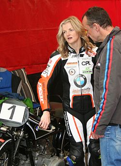 Tall slim woman with long blonde hair wearing multi coloured racing leathers standing next to an historic BMW motorcyle in the company of an older man