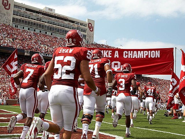 Football players run onto a football field in two rows under a crimson banner.