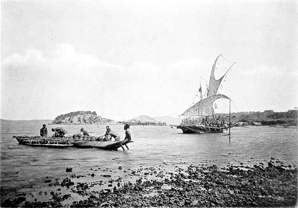 Black and white photograph of six men loading a boat on a beach, with a sailing boat just off shore.