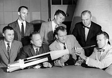 Sept hommes, quatre assis et trois debout, regardent une maquette de fusée.