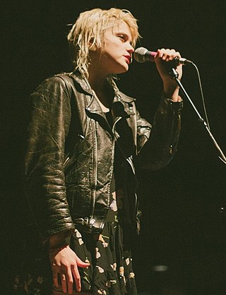 A light-skinned blonde woman wearing a leather jacket is seen singing into her microphone