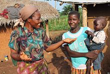 Angélique Namaika. Afrikanische Frau mit bunte Kleidern und einem braunen Band um den Kopf im Gespräch mit einer Person, die ein Kind auf dem Arm hält (Vertriebene). In einem Dorf aus schilfgedeckten Lehmhütten.