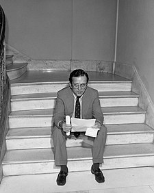 Stevens in 1977 as Assistant Minority Leader. He is seated on some steps, looking up, with black hair and glasses, wearing a Senator's usual suit and tie. He is holding a sheet of paper.