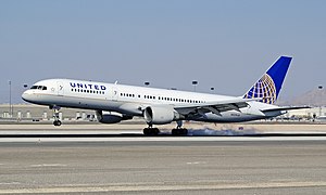 A mostly white Boeing 757 with blue and yellow trim preparing for landing against a blue sky. Landing gear and flaps are fully extended in final approach configuration.