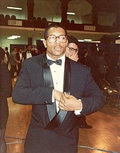 A man wearing a tuxedo and eyeglasses; other people can be seen in the background, along with folding chairs.
