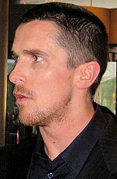 Profilový snímek muže, který se dívá nalevo.  Má kozí bradku a strniště a na sobě má černou společenskou košili s límečkem a černým blejzrem.