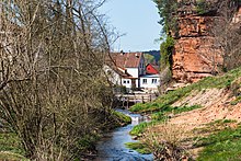 Blick von einer Brücke auf den Kondeler Bach. Rechts davon befindet sich eine Felswand. Links davon Gestrüpp und Häuser. Auch sieht man eine weitere Brücke über den Bach.