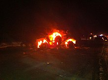 At night, armoured car sitting engulfed in flames in the middle of a road