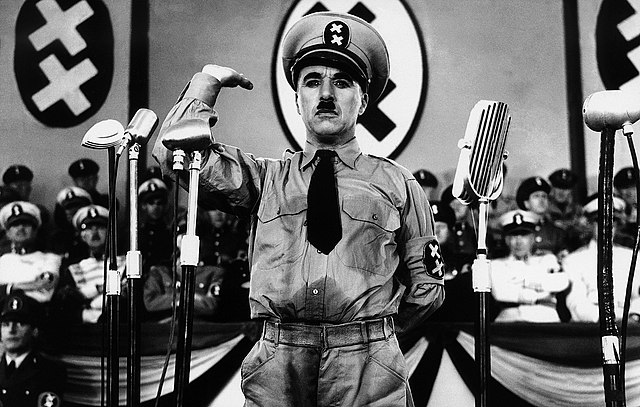 Photographie en noir et blanc tirée d'un film, où le personnage principal est habillé en dictateur levant la main dans une parodie de salut nazi.